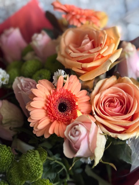 A close-up view of a floral bouquet featuring peach and pink roses, arranged alongside a coral pink gerbera daisy with a dark center, and small green berries, all wrapped in soft pink paper with a sub