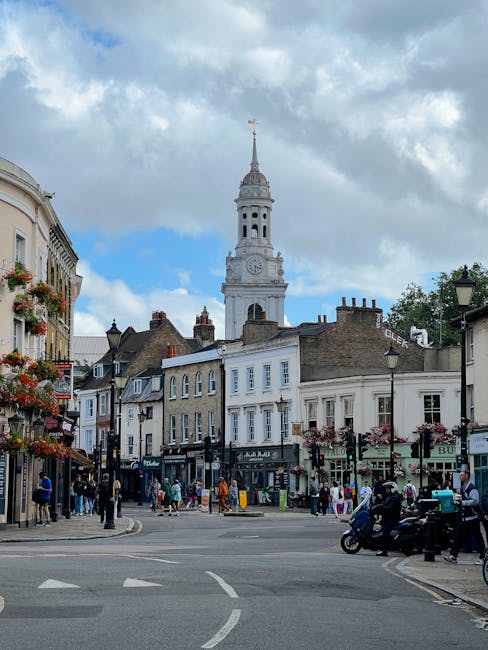 The image depicts a lively town square in Beckenham, with a prominent historic clock tower rising above a row of traditional Georgian-style buildings. The street is bustling with pedestrians, some bro