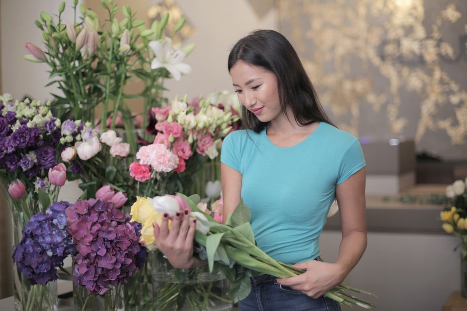 A woman in a light blue T-shirt, inside a flower shop, carefully arranging a bouquet of fresh flowers, including white lilies, pink roses, and yellow tulips. She is holding the bouquet with both hands
