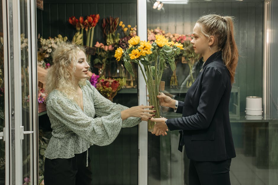 A florist with curly blonde hair dressed in a light-colored, patterned blouse hands a bouquet of bright yellow sunflowers wrapped in clear cellophane to a young woman with long brown hair styled in a 