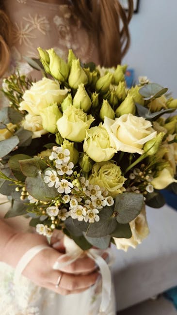 A floral bouquet featuring cream-colored roses and light green buds, accented with small white daisy-like flowers and sprigs of eucalyptus leaves. The arrangement is held by a person wearing a cream-c