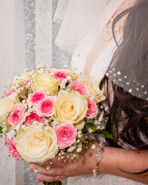 A bride holding a bouquet of fresh flowers comprising large ivory roses and smaller pink roses, accented with delicate white baby's breath and green foliage. The bouquet is tightly arranged in a round