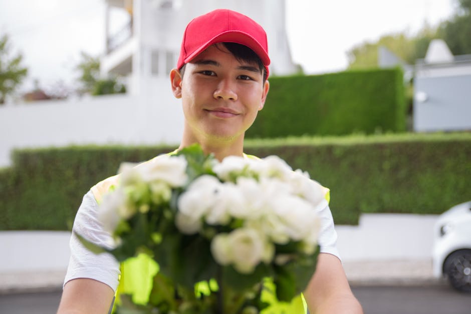 A delivery person wearing a red cap and a high-visibility vest holds out a bouquet of fresh white roses and green foliage towards the camera. The flowers are arranged in a compact, rounded floral arra